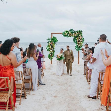 Impressive Punta Cana Exterior foto The photo depicts a wedding ceremony on a beach. In the center, there is an altar decorated with greenery and flowers. A couple is walking down the aisle, with guests on either side capturing the moment on their phones. The attendees are dressed in a