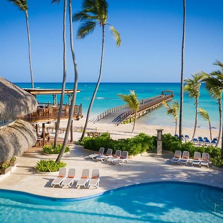 Impressive Punta Cana Exterior foto The photo depicts a serene tropical scene featuring a swimming pool in the foreground, surrounded by lounge chairs. In the background, there is a beautiful beach with clear turquoise waters and a wooden pier extending out into the ocean. Palm trees f