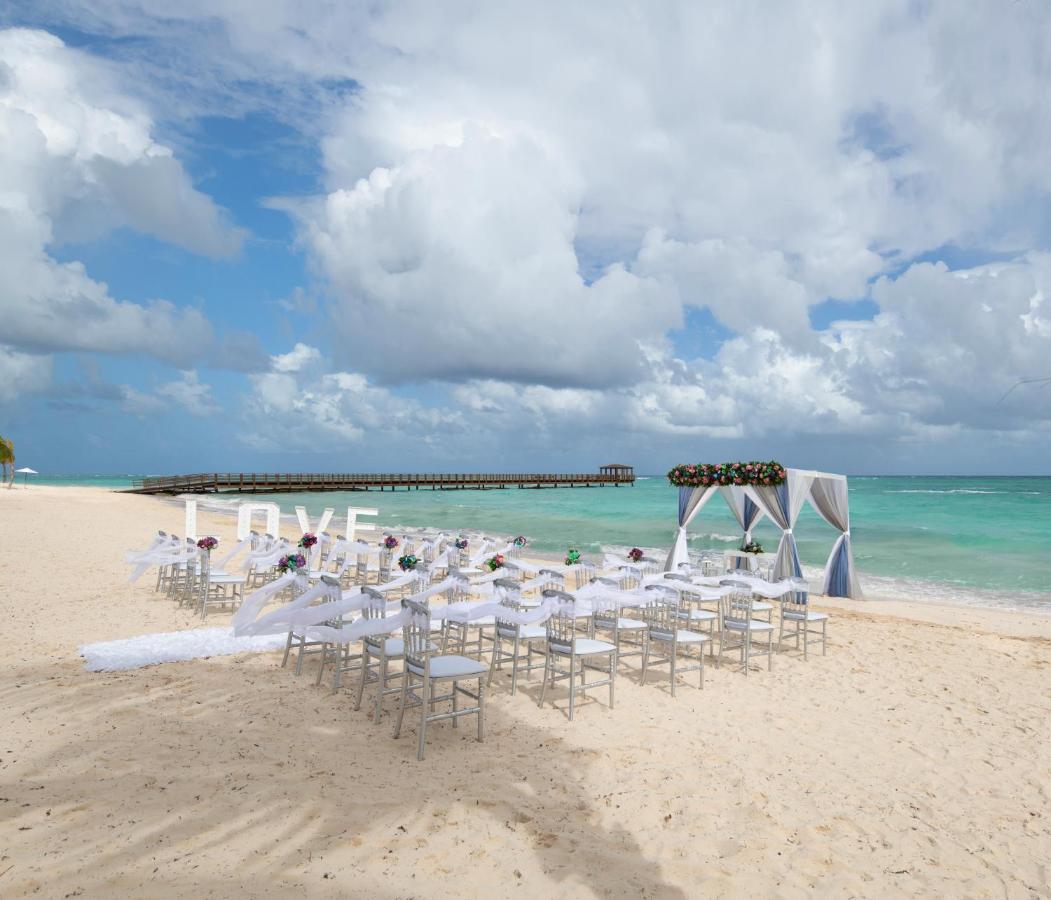 Impressive Punta Cana Exterior foto The photo shows a beach setting that appears to be prepared for a wedding ceremony. There are rows of white chairs arranged on the sand facing a decorated archway. The arch is draped in white fabric and adorned with floral elements. In the background