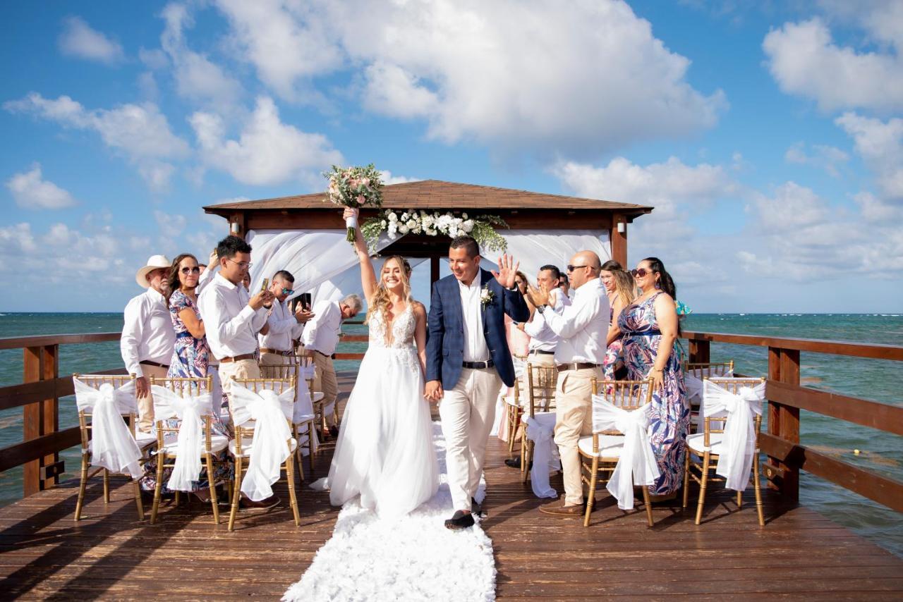 Impressive Punta Cana Exterior foto The photo shows a wedding ceremony taking place on a wooden pier over water. A bride and groom are at the center, with the bride holding her bouquet up in celebration. They are surrounded by a group of guests, all dressed in formal attire, some clapp