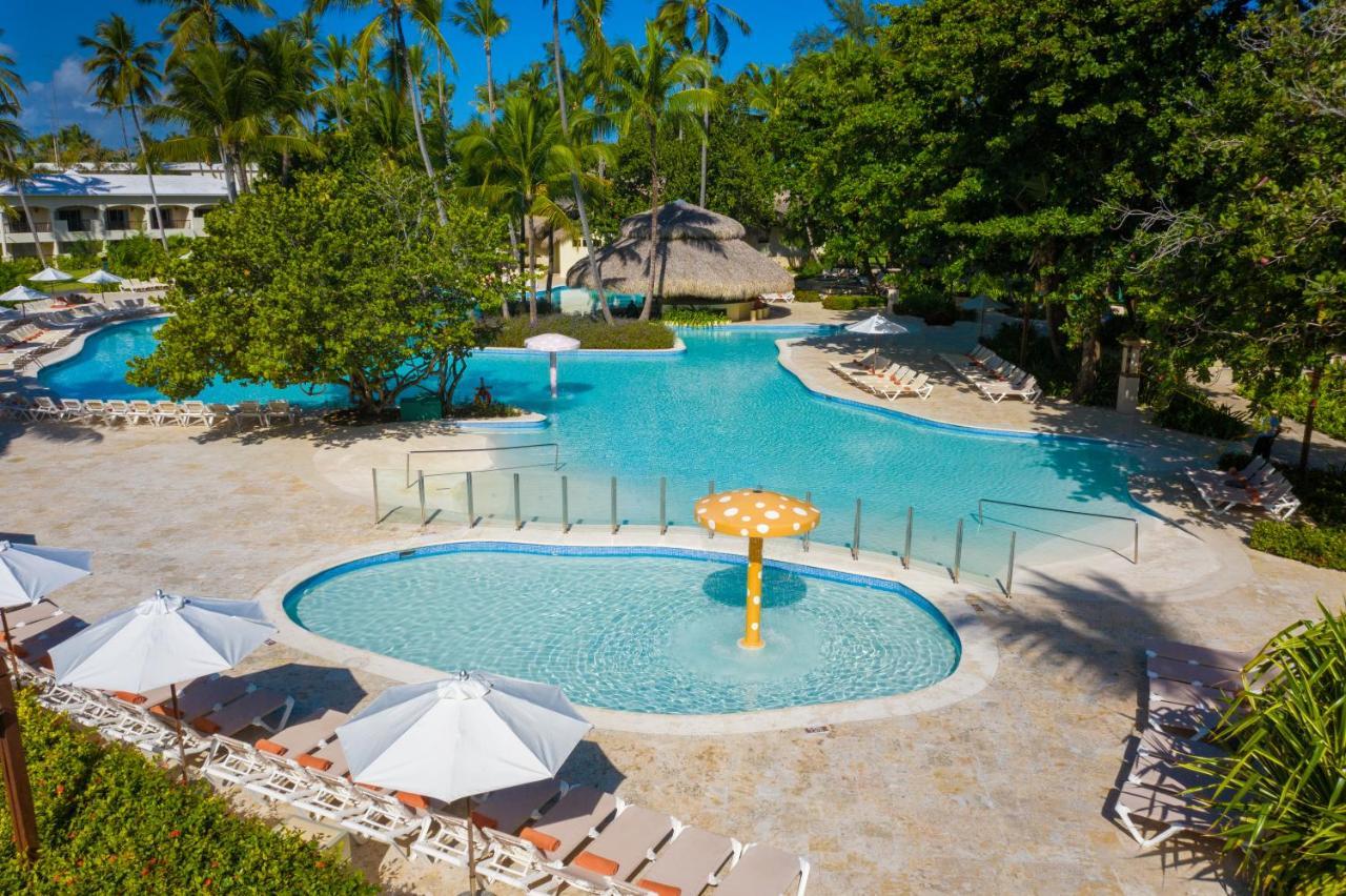 Impressive Punta Cana Exterior foto The photo shows a vibrant outdoor pool area surrounded by lush greenery. There are two distinct pool sections visible: a larger main pool with a clear blue water and a smaller, circular pool that features a playful mushroom-shaped fountain. Sun loung