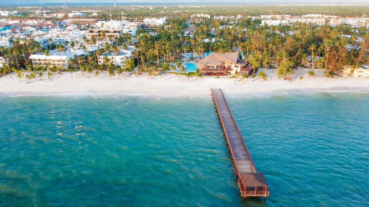 Impressive Punta Cana Exterior foto The photo shows a picturesque beach scene viewed from above. In the foreground, there is a wooden pier extending over clear turquoise water. The pier leads towards a coastal area with a sandy beach lined by lush palm trees. In the background, several