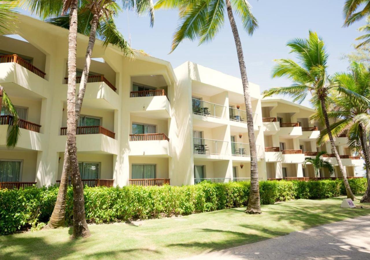 Impressive Punta Cana Exterior foto The photo shows a beachfront hotel or resort building with multiple levels. The structure is light-colored with balconies that overlook a manicured garden area. Tall palm trees and lush green vegetation surround the building, suggesting a tropical lo