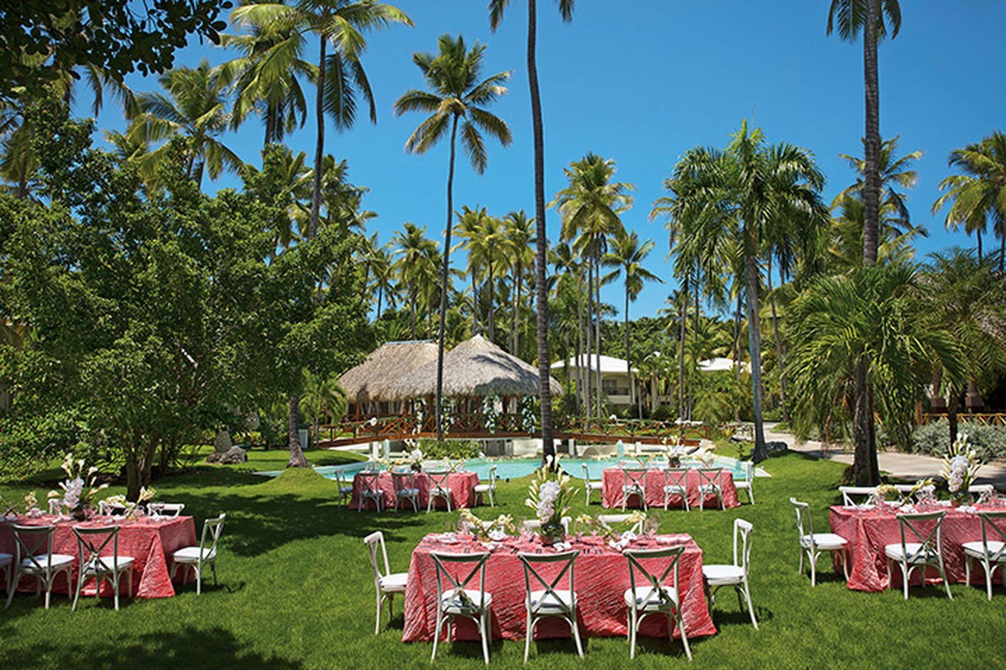 Impressive Punta Cana Exterior foto The photo depicts a beautiful outdoor setting, likely for an event or celebration. There are several tables arranged on a lush green lawn, each dressed with pink tablecloths. White chairs surround the tables, creating an inviting atmosphere. In the b
