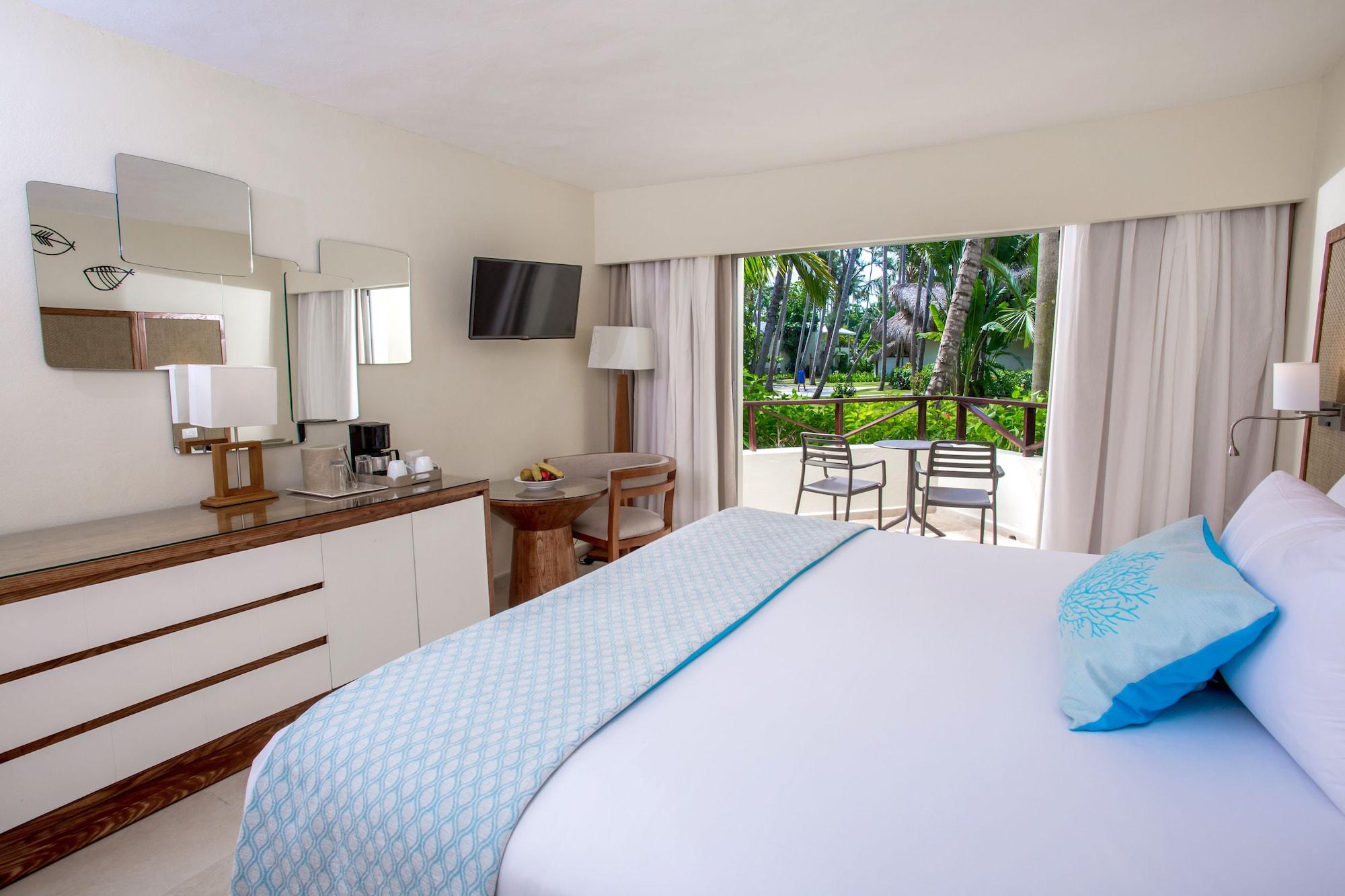 Impressive Punta Cana Exterior foto The photo shows a well-decorated hotel room featuring a large bed with a light blue throw blanket. There is a bedside table and a dresser with a mirror, as well as a television mounted on the wall. Natural light comes in through a large window that o