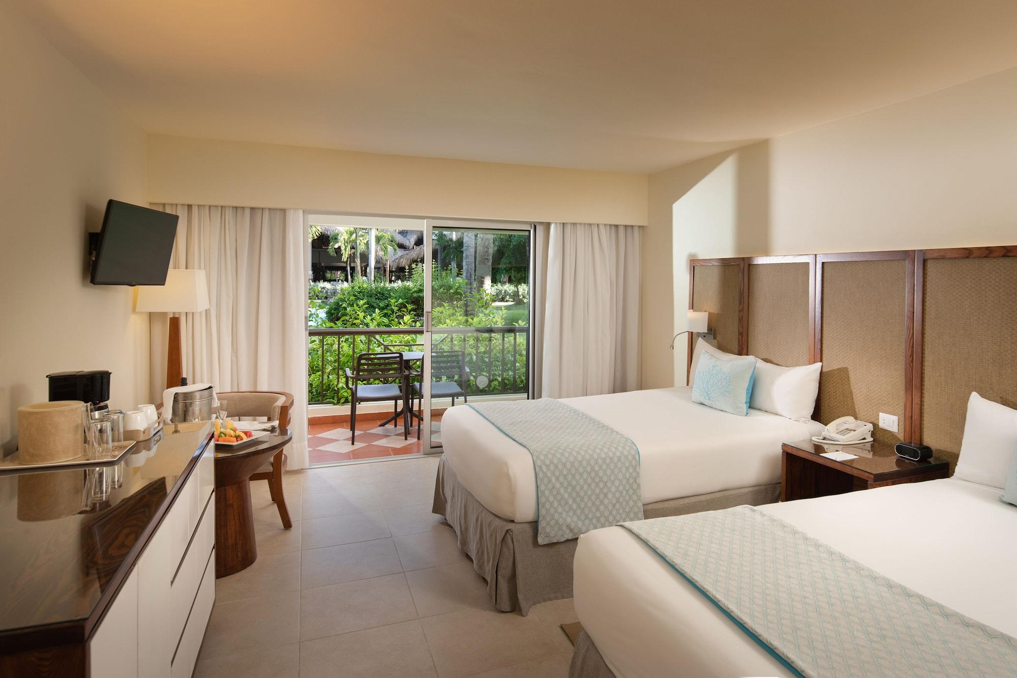 Impressive Punta Cana Exterior foto The photo shows a hotel room featuring two beds with light-colored linens and decorative pillows. There is a modern armchair beside a small table near a large window that offers a view of an outdoor garden area. A flat-screen TV is mounted on the wal