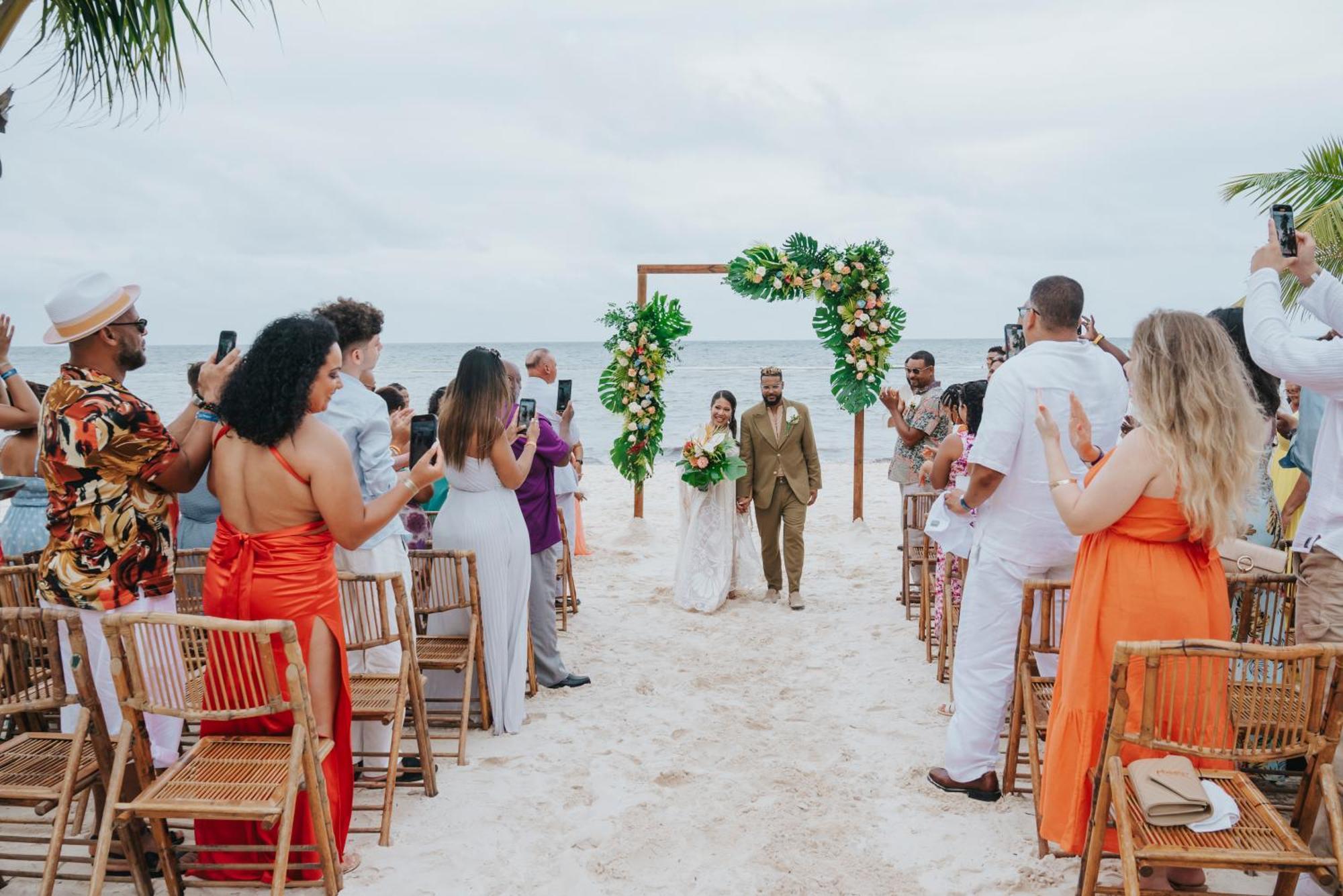 Impressive Punta Cana Exterior foto The photo depicts a wedding ceremony on a beach. In the center, there is an altar decorated with greenery and flowers. A couple is walking down the aisle, with guests on either side capturing the moment on their phones. The attendees are dressed in a