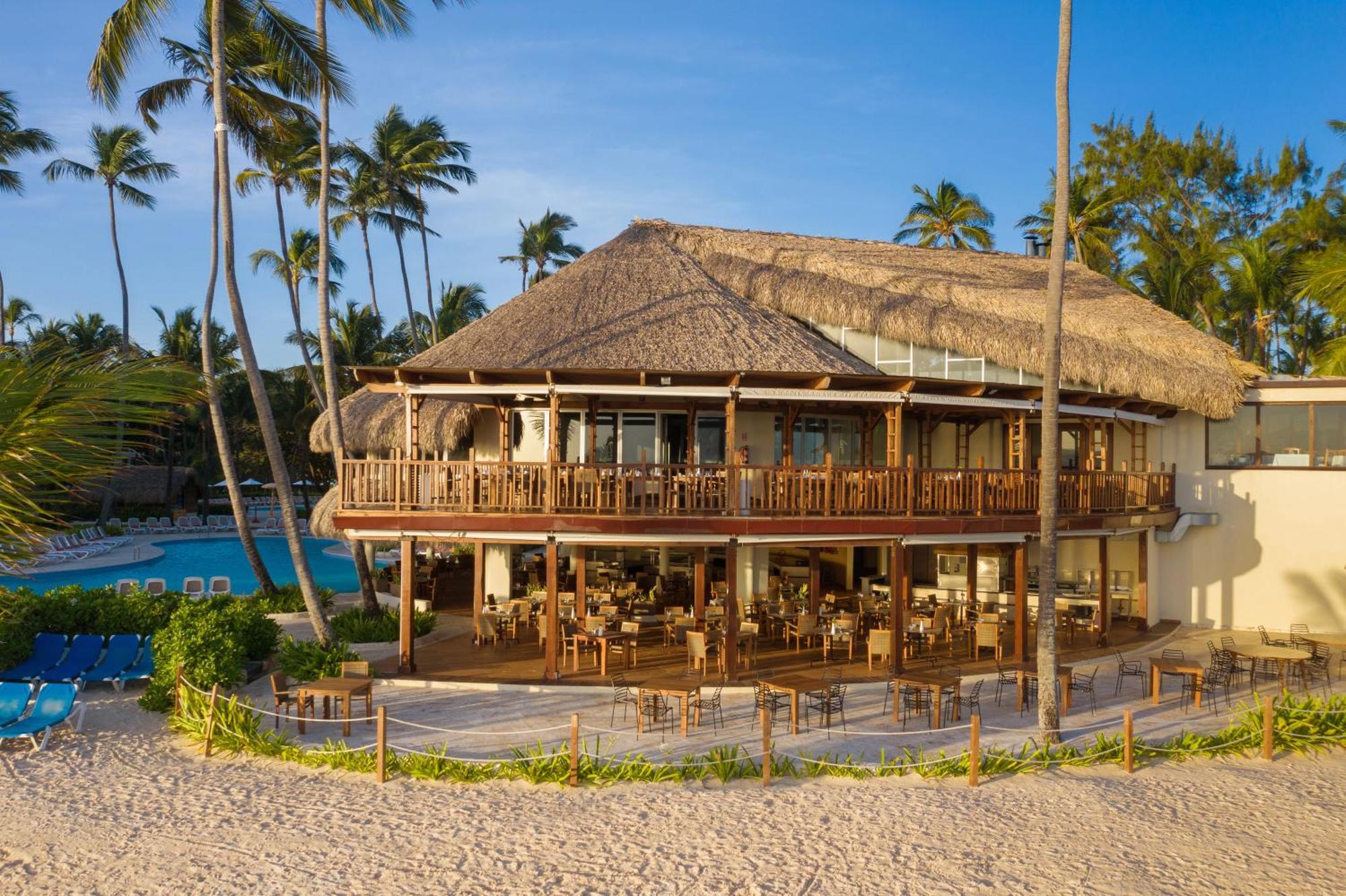 Impressive Punta Cana Exterior foto The photo shows a tropical beachfront building, likely a restaurant or resort, featuring a large deck with wooden railings and outdoor seating. The structure has a thatched roof and multiple large windows, allowing for a view of the surrounding area.