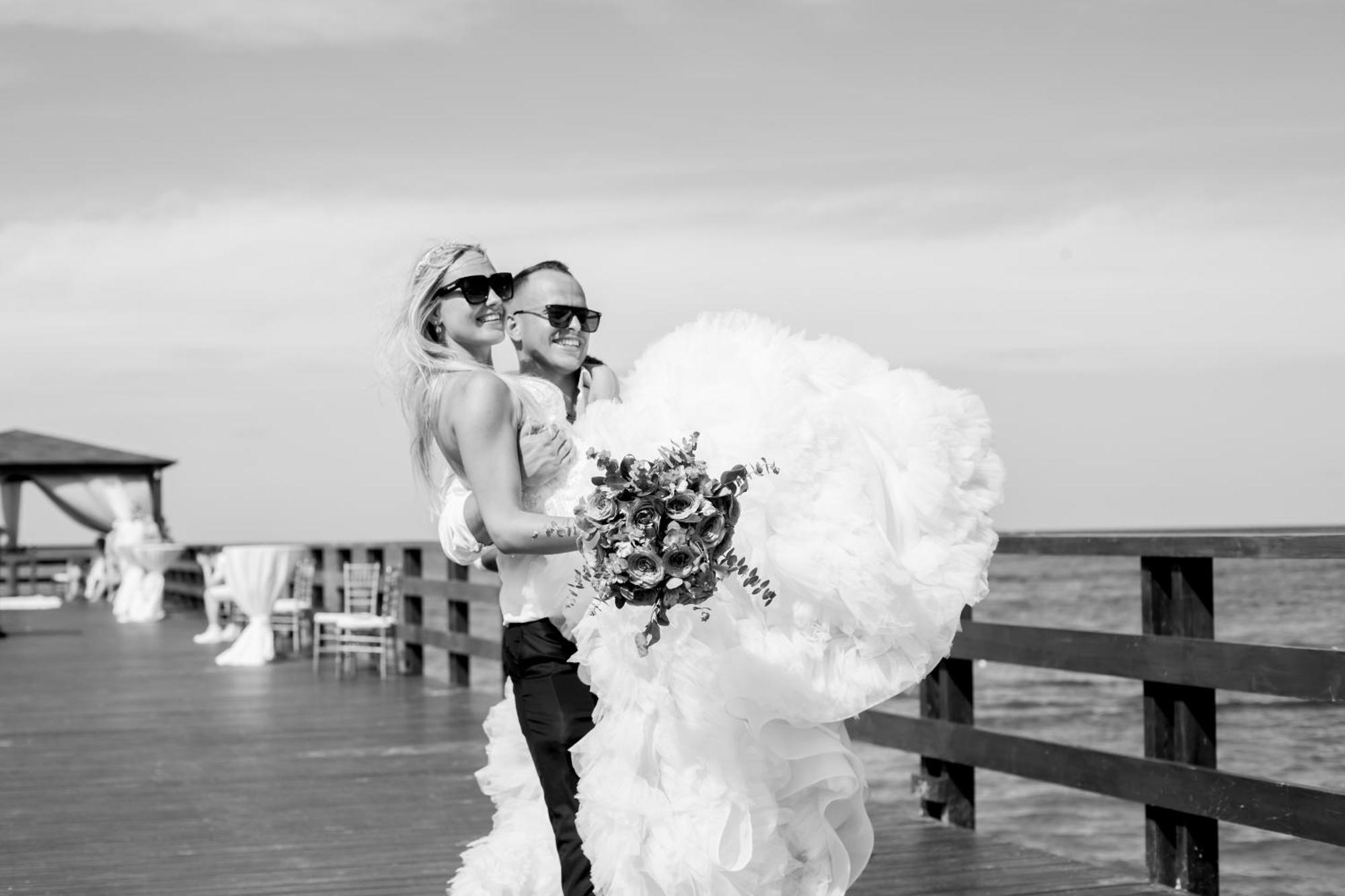Impressive Punta Cana Exterior foto The photo appears to depict a couple celebrating their wedding. They are standing on a wooden pier by the water, with the ocean in the background. The scene is in black and white, which adds a classic touch. The bride is wearing a dramatic, fluffy we