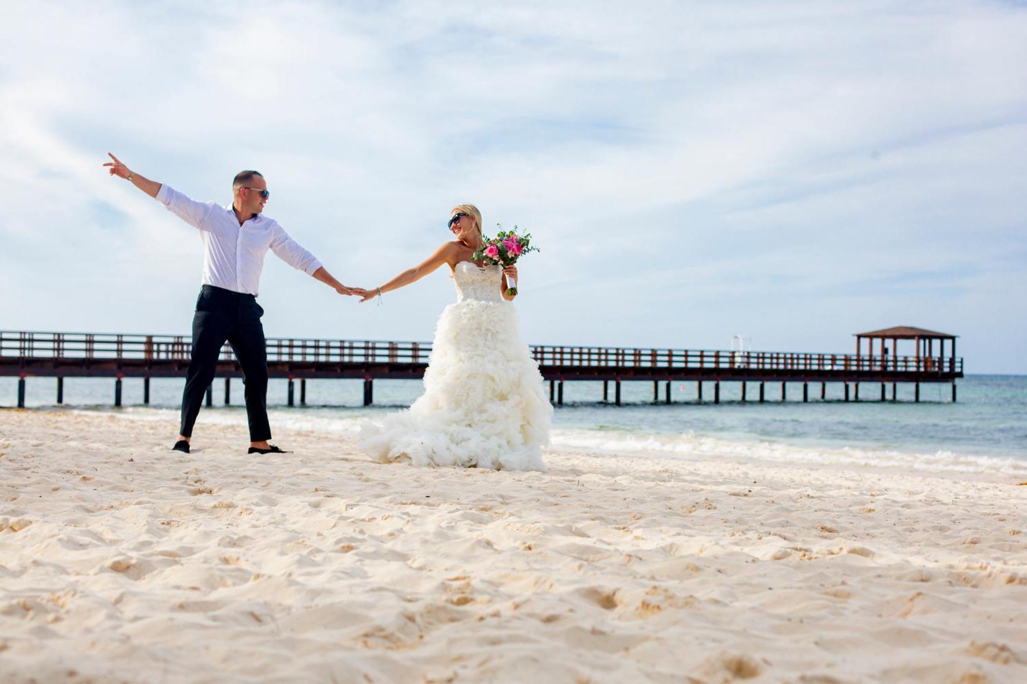 Impressive Punta Cana Exterior foto The photo depicts a joyful couple on a beach. The bride wears a beautiful white wedding gown and holds a bouquet of flowers, while the groom is dressed in a light shirt and dark pants. They are standing on the sandy shore, holding hands and smiling a