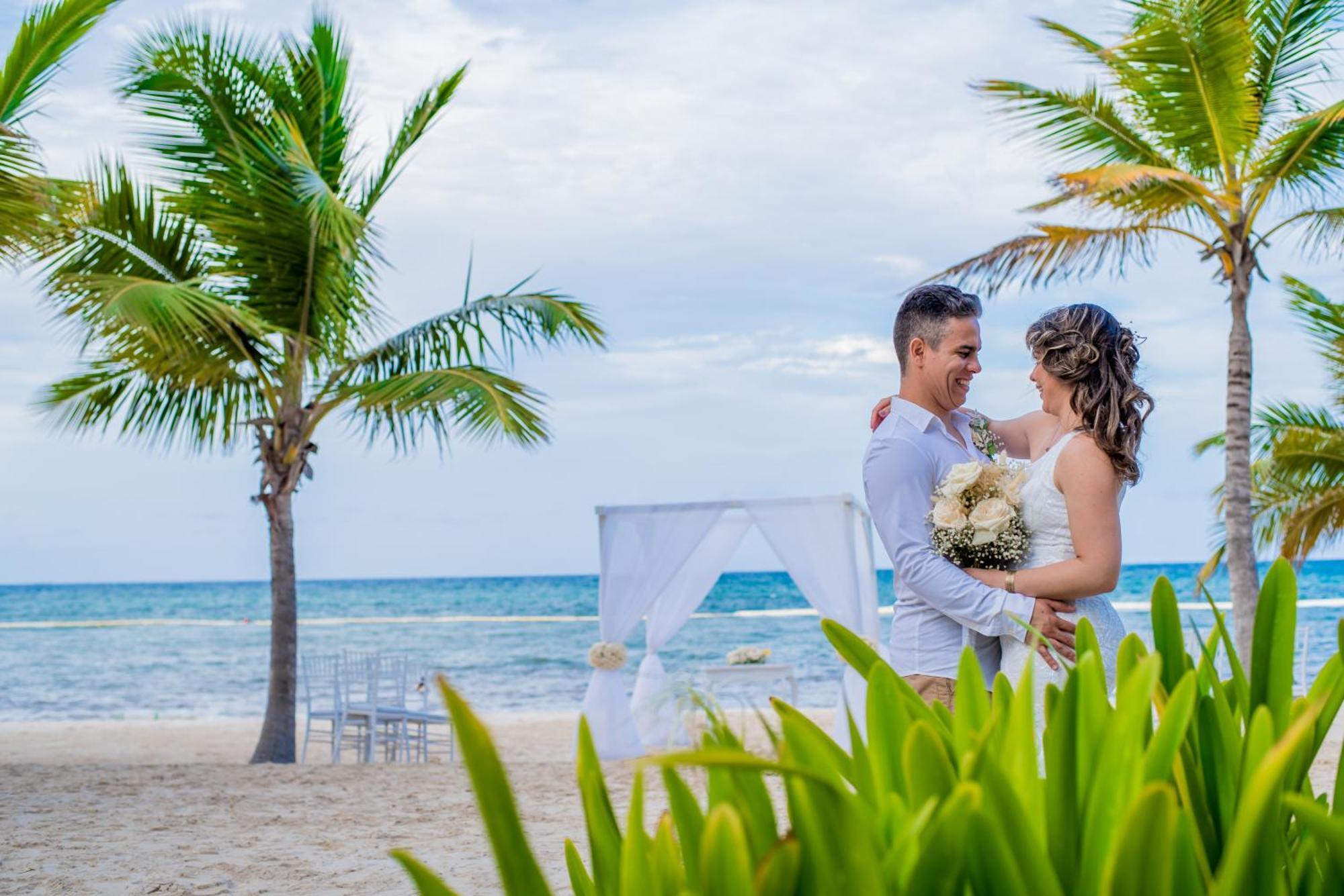 Impressive Punta Cana Exterior foto The photo depicts a couple embracing on a beach. They appear to be celebrating their wedding, as evidenced by the woman's bouquet. The scenic background features palm trees and the ocean, creating a romantic atmosphere. A white wedding canopy is set 