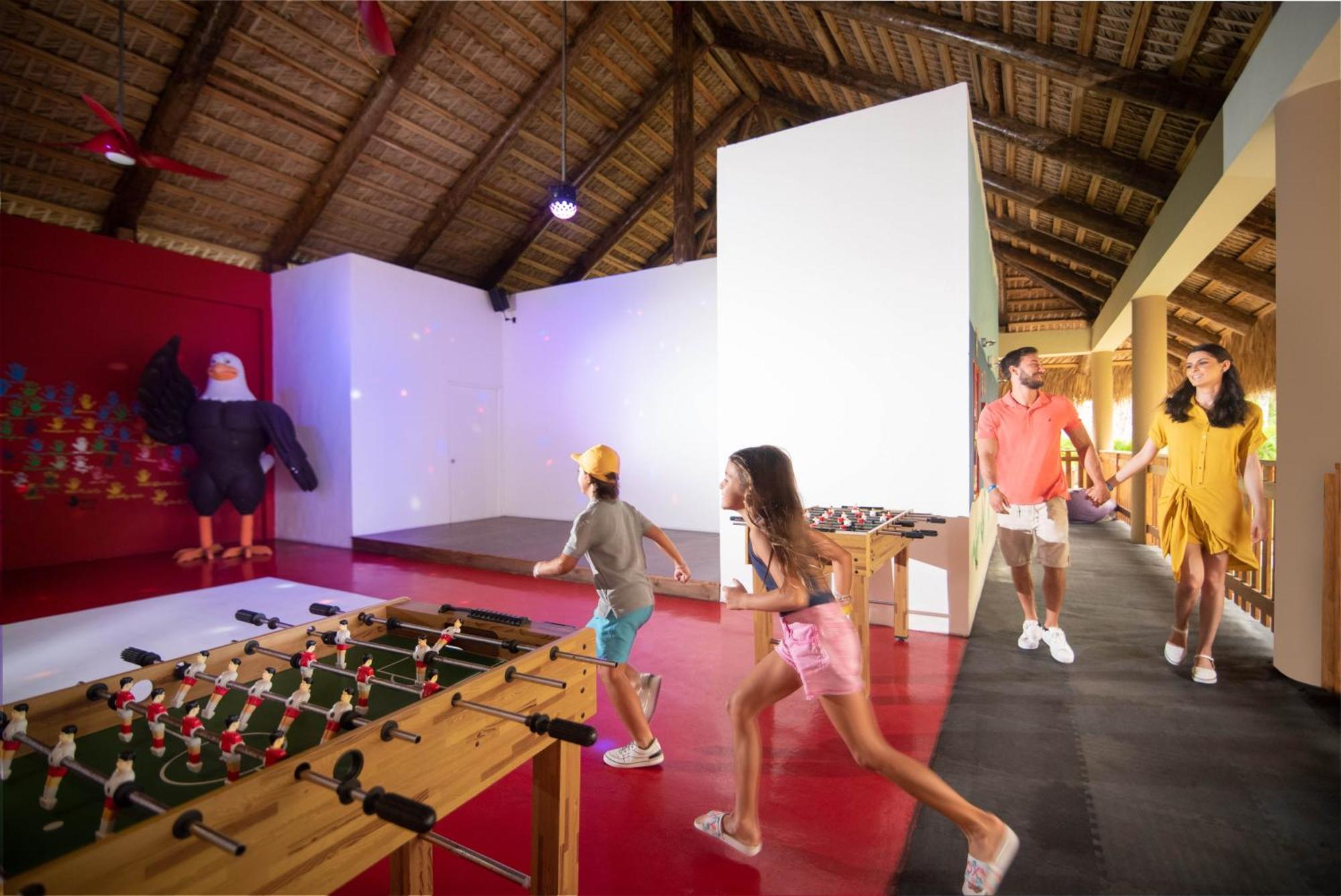 Impressive Punta Cana Exterior foto The photo shows an indoor recreational space with a thatched roof. In the foreground, there is a foosball table with figures attached to it, where two children are playing or running towards it. A man and a woman, likely parents, are walking together