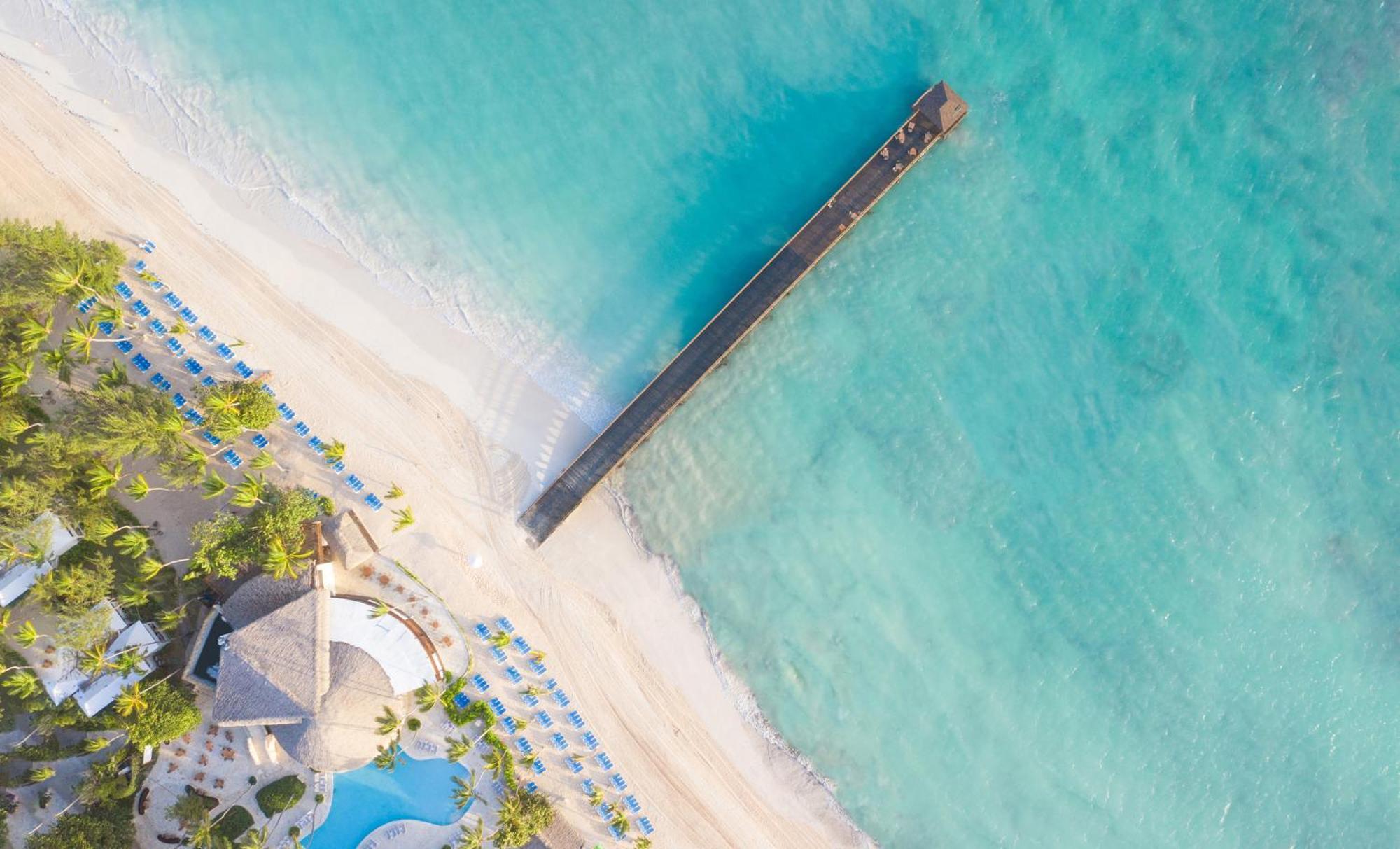 Impressive Punta Cana Exterior foto The photo shows an aerial view of a beautiful beach scene. There is a long wooden pier extending over clear turquoise waters, leading out from the sandy shore. The beach is lined with umbrellas or lounge chairs, and there is a large building with a t