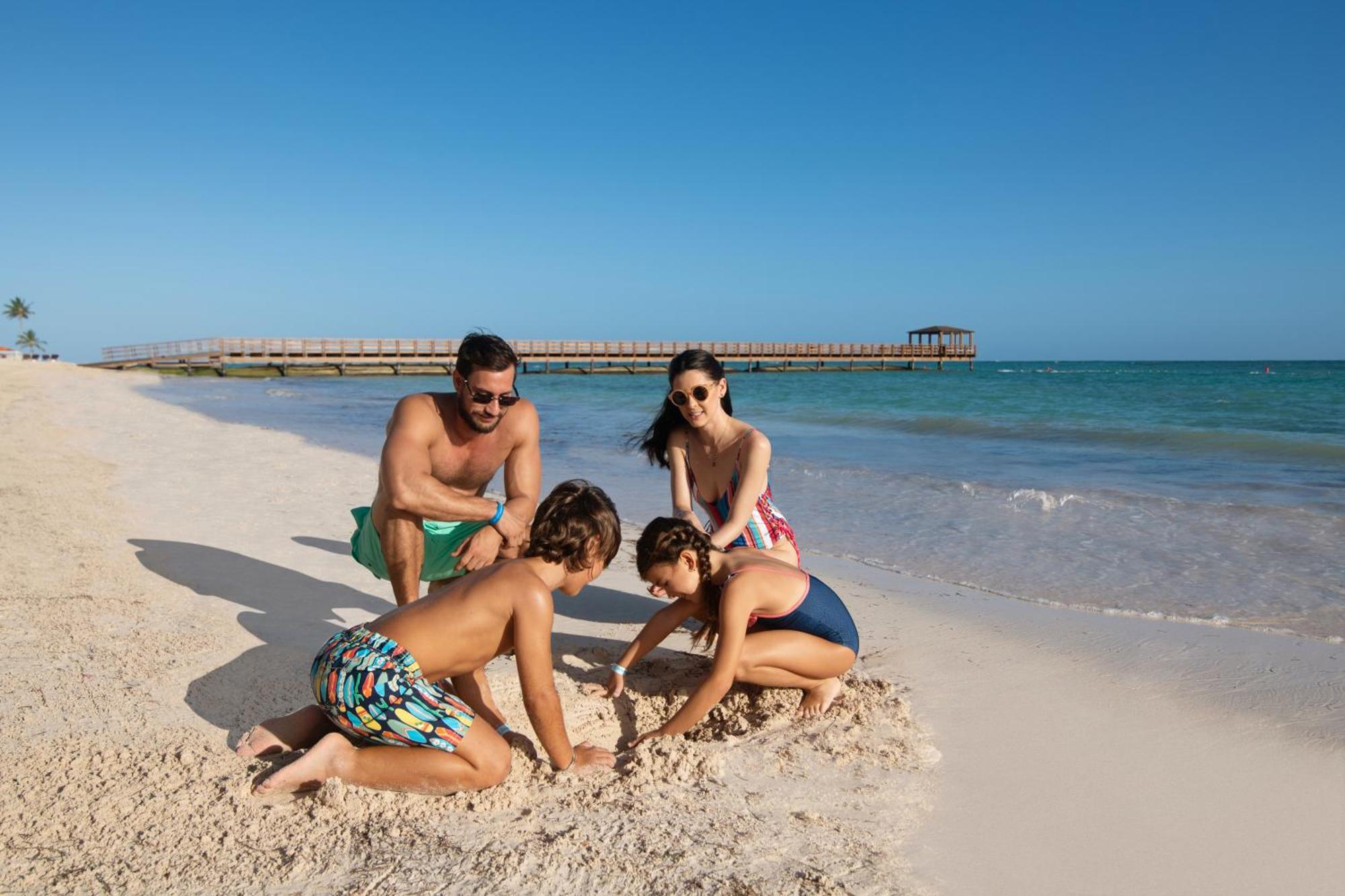 Impressive Punta Cana Exterior foto The photo depicts a family enjoying a sunny day at the beach. In the foreground, two children are playing in the sand, focused on digging or building something. One child is kneeling, while the other is sitting nearby. An adult male is watching them 