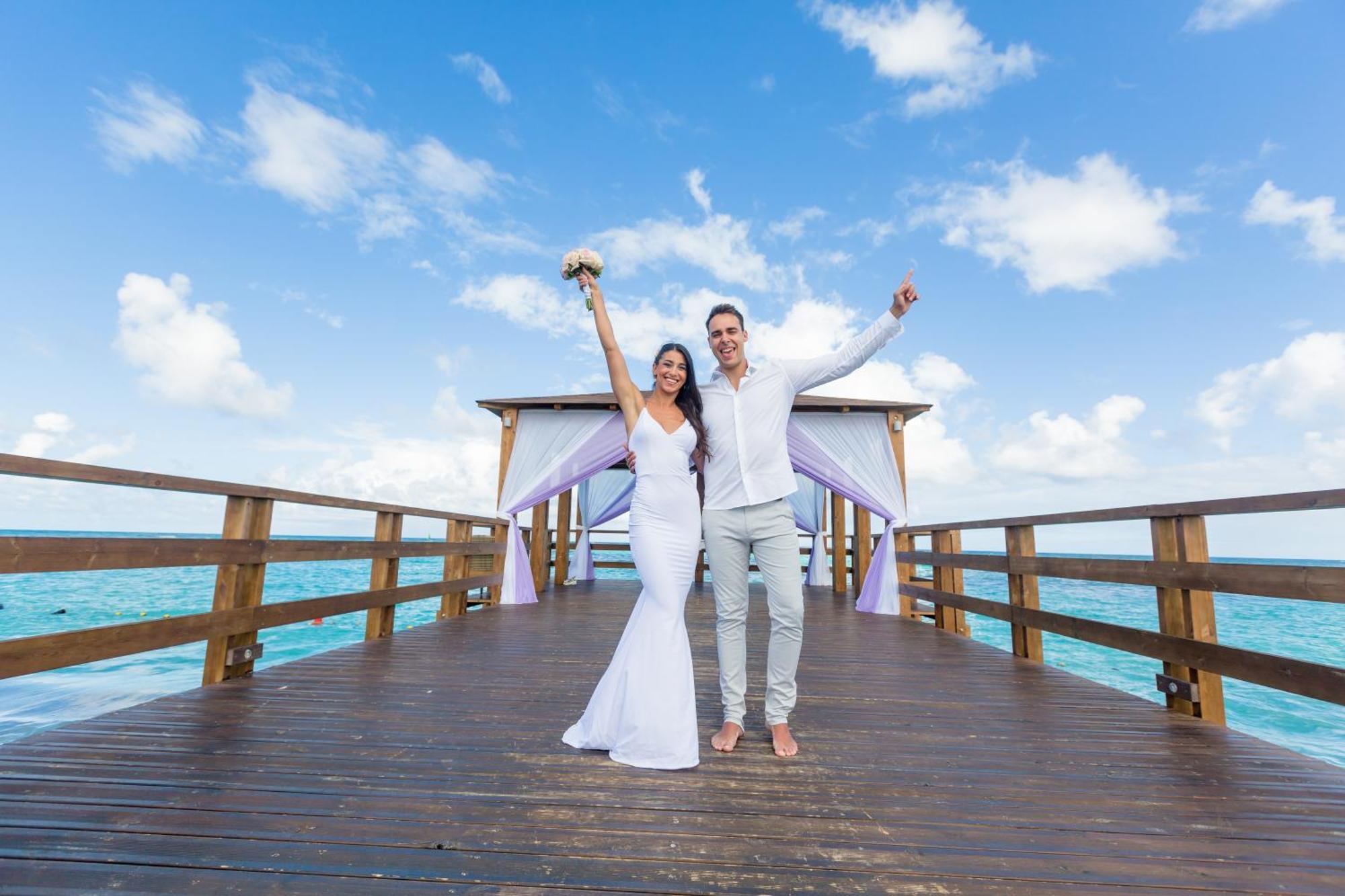 Impressive Punta Cana Exterior foto The photo shows a joyful couple standing on a wooden pier or deck over the water. The woman is wearing a beautiful white wedding dress and is holding a bouquet of flowers, while the man is dressed in light-colored attire, likely suitable for a beach 