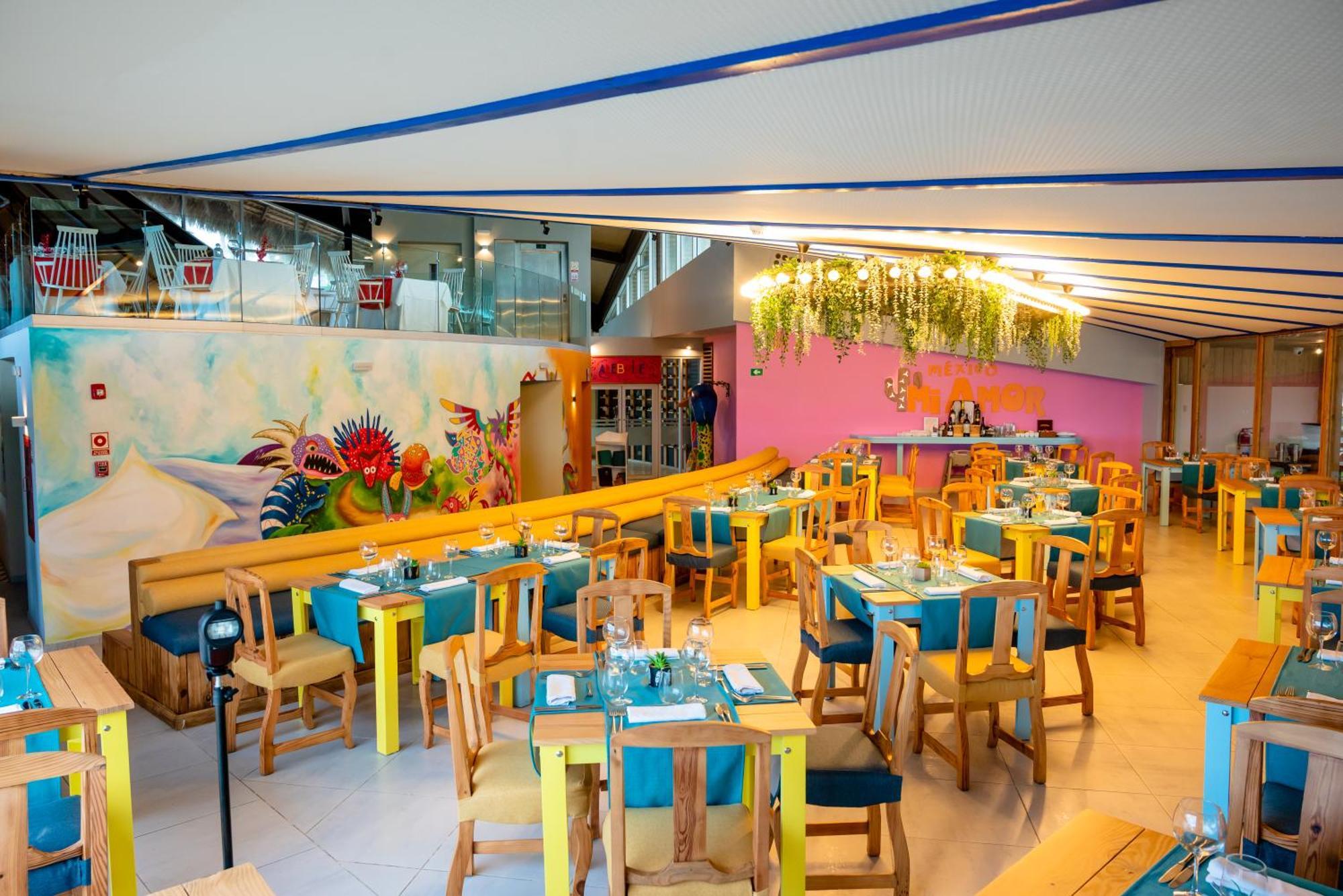 Impressive Punta Cana Exterior foto The photo shows a vibrant restaurant interior. It features wooden tables and chairs, painted in bright, cheerful colors like yellow and turquoise. The tables are set with utensils and blue tablecloths. The walls are decorated with colorful murals and