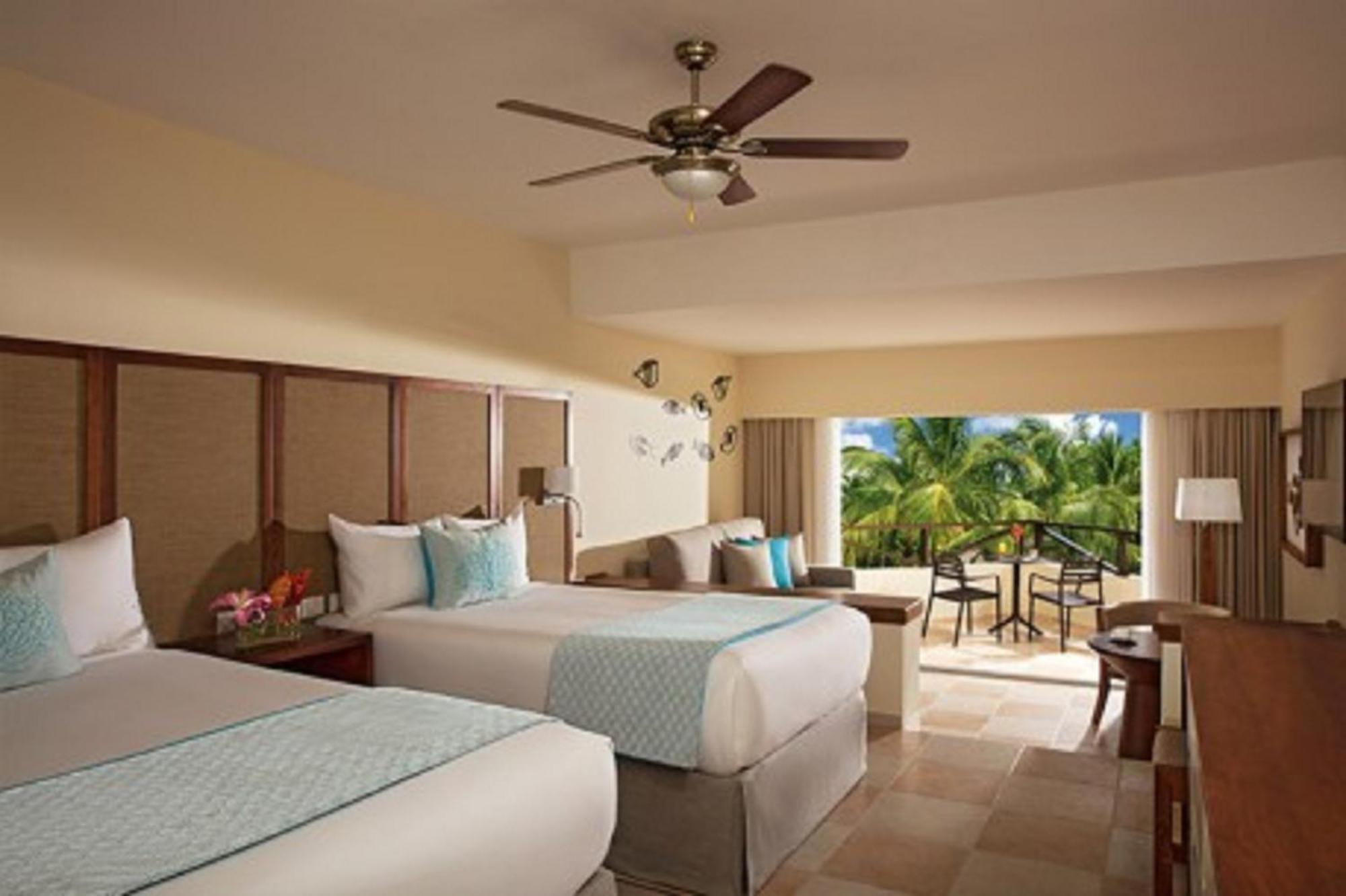Impressive Punta Cana Exterior foto The photo shows a hotel room with two beds, decorated in a light, modern style. There is a ceiling fan above and large windows that open to a balcony. The room features soft colors, with a turquoise accent on the bedding, and has a small seating area