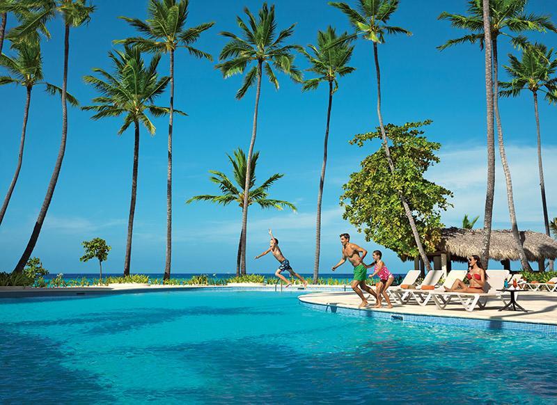 Impressive Punta Cana Exterior foto The photo showcases a vibrant tropical scene featuring a swimming pool surrounded by tall palm trees. In the foreground, several people are enjoying themselves, with one person jumping into the pool. Others are relaxing on lounge chairs by the poolsi