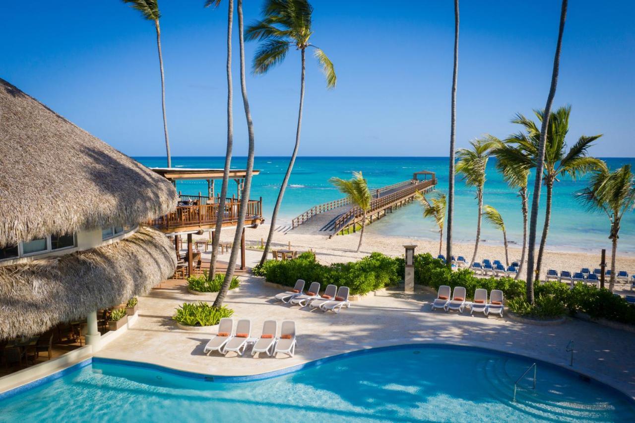 Impressive Punta Cana Exterior foto The photo depicts a serene tropical scene featuring a swimming pool in the foreground, surrounded by lounge chairs. In the background, there is a beautiful beach with clear turquoise waters and a wooden pier extending out into the ocean. Palm trees f