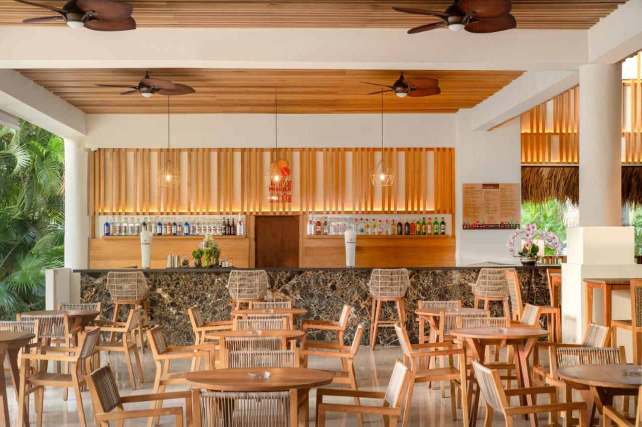 Impressive Punta Cana Exterior foto The photo shows a modern and inviting bar or restaurant interior. It features a spacious layout with wooden flooring and ceiling, complemented by ceiling fans. The bar area is prominently displayed in the background, showcasing a variety of bottles w