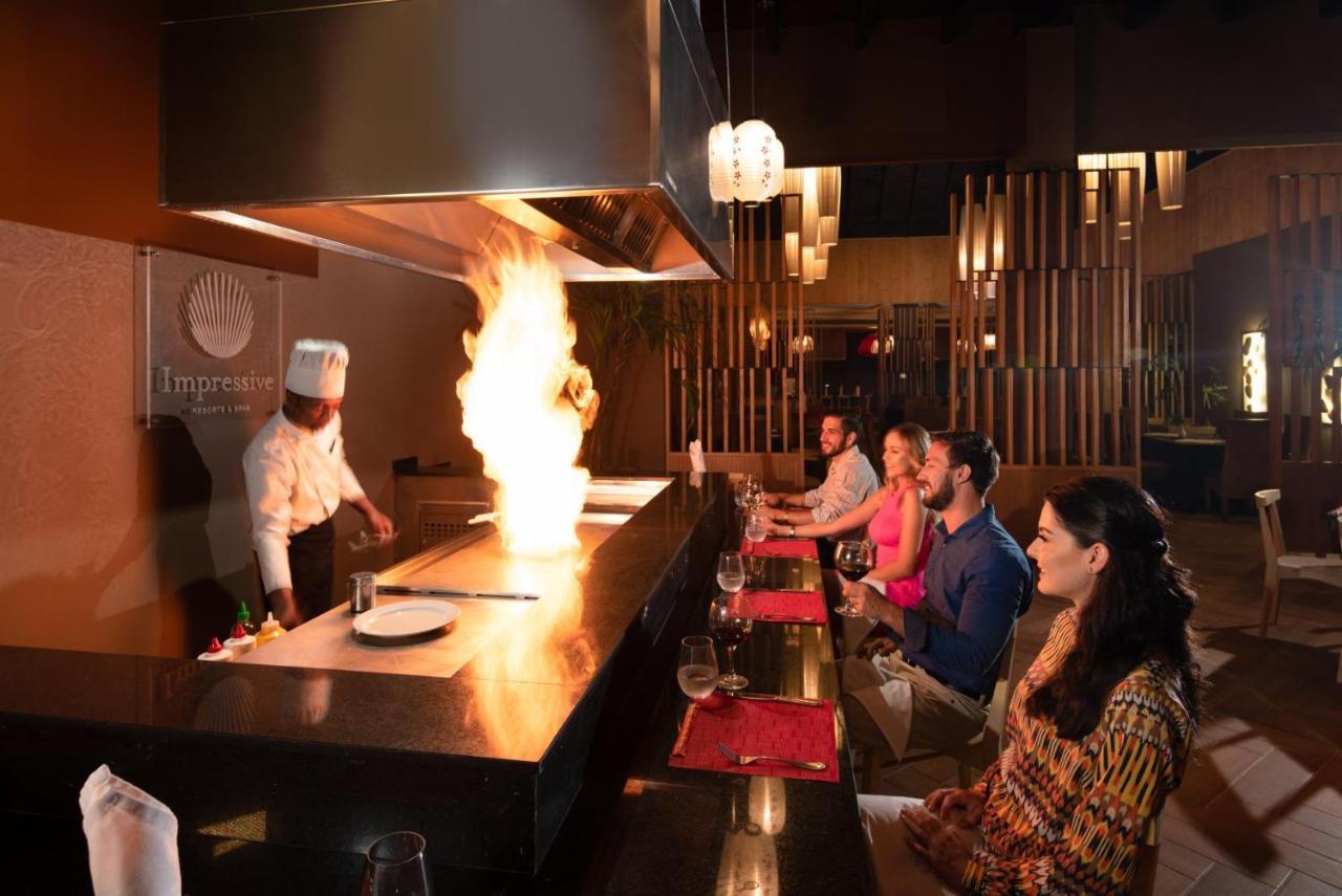 Impressive Punta Cana Exterior foto The photo shows a dining scene in a restaurant setting. A chef in a white uniform is performing a cooking demonstration at a teppanyaki table, creating a large fire as part of the cooking process. In front of the chef, several diners are sitting at t