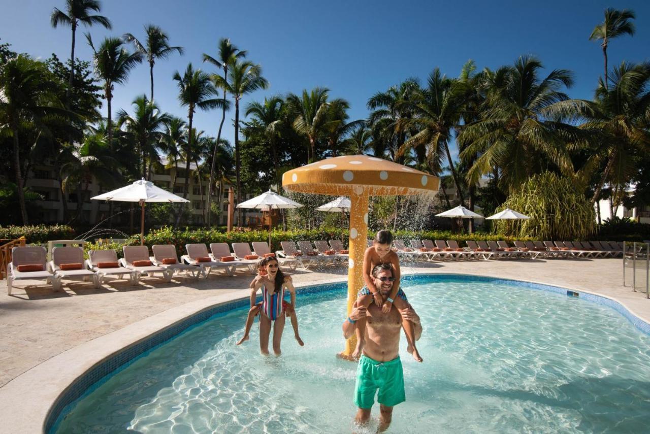 Impressive Punta Cana Exterior foto The photo shows a lively pool scene under a clear blue sky. In the foreground, there are two children playing; one is sitting on the shoulders of an adult. They are enjoying the water and the sunny atmosphere. Nearby, there is a water feature resembl